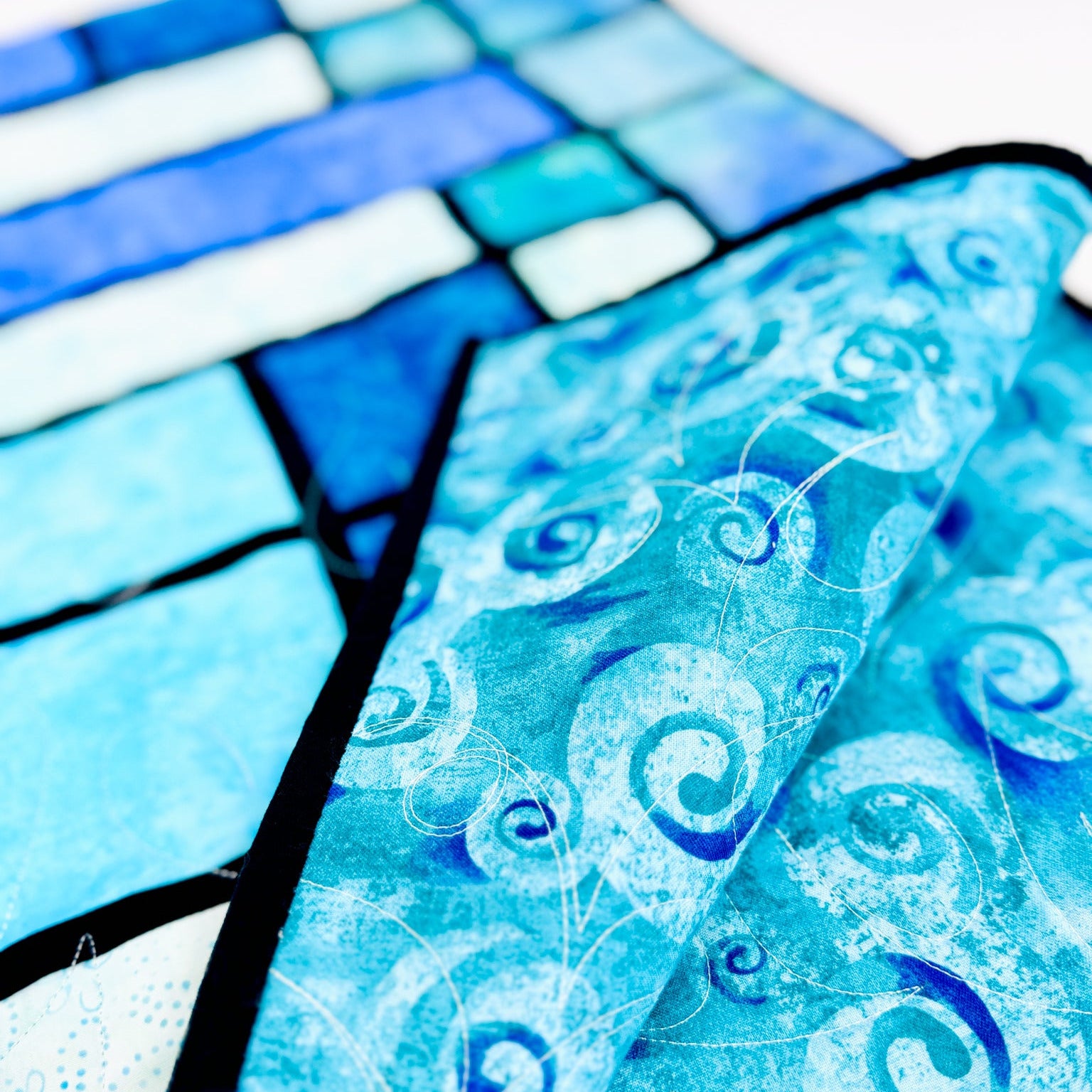 A close-up of a blue and turquoise patterned fabric item, likely a quilt or textile panel, with swirling wave-like motifs and black stitched borders, shown against a white background.
