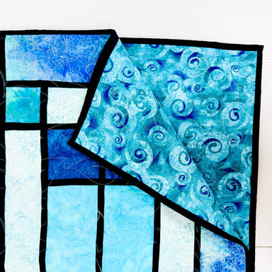 A close-up of a quilted textile featuring geometric panels and a swirled pattern in shades of blue, aqua, and white, separated by black quilting lines.