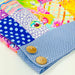A close-up of a colorful patchwork quilted fabric item, likely a blanket or quilt, featuring a blue gingham folded corner with two wooden buttons.