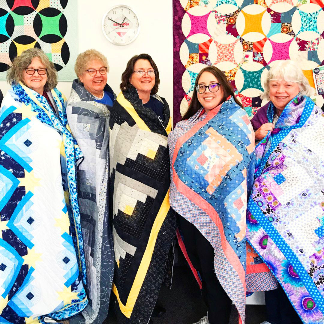 Five women standing together, each holding a colorful quilt in front of a wall with more quilts displayed.