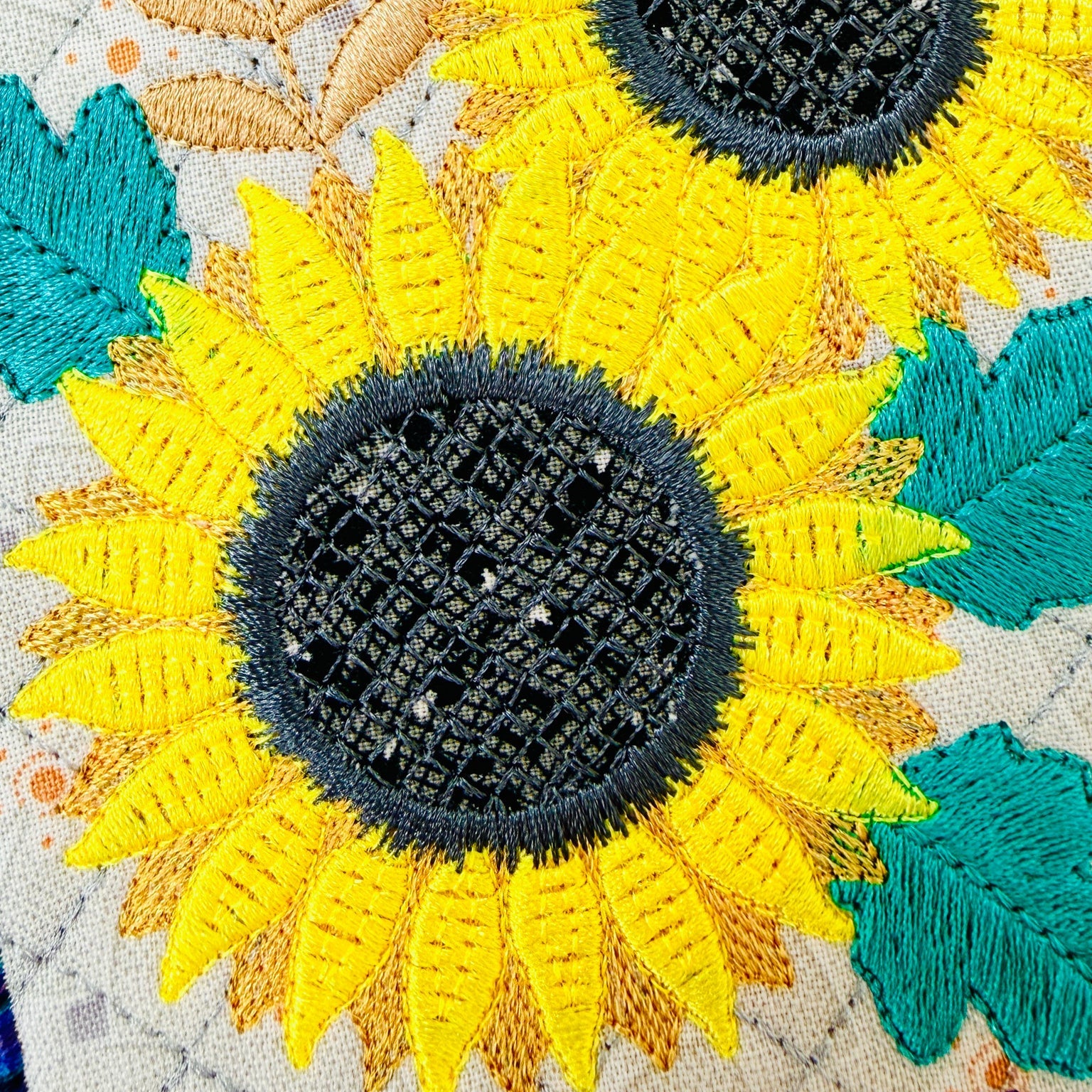 Close-up of embroidered sunflowers with bright yellow petals, dark gray centers, teal leaves, and peach stems on a light fabric background.