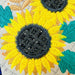 Close-up of embroidered sunflowers with bright yellow petals, dark gray centers, teal leaves, and peach stems on a light fabric background.