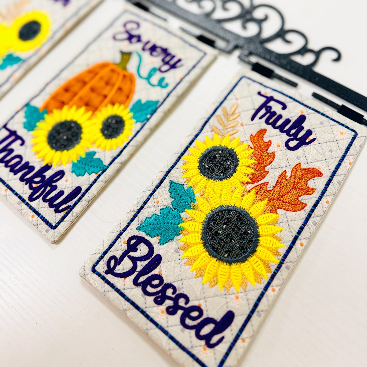 Three decorative embroidered fabric banners hanging from a black metal scroll bar against a white background. The banners feature autumn-themed embroidery including sunflowers, pumpkins, leaves, and phrases like 'So very nice,' 'Fruity Blessed,' and 'Thankful.