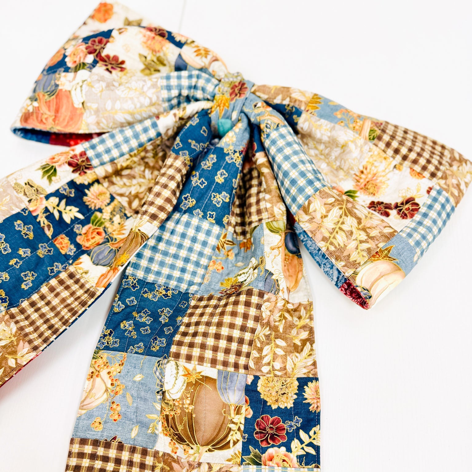 A large fabric bow with a patchwork print featuring blue gingham, brown gingham, floral motifs, and a central gold-toned embellishment, photographed on a white background.