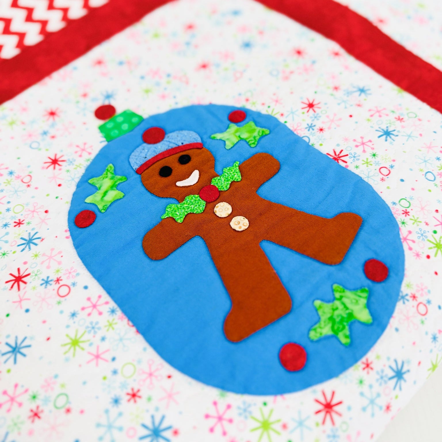 A close-up of a colorful quilted fabric featuring a gingerbread man applique inside a blue circle, surrounded by a white fabric with multicolored snowflake patterns and a red chevron border.