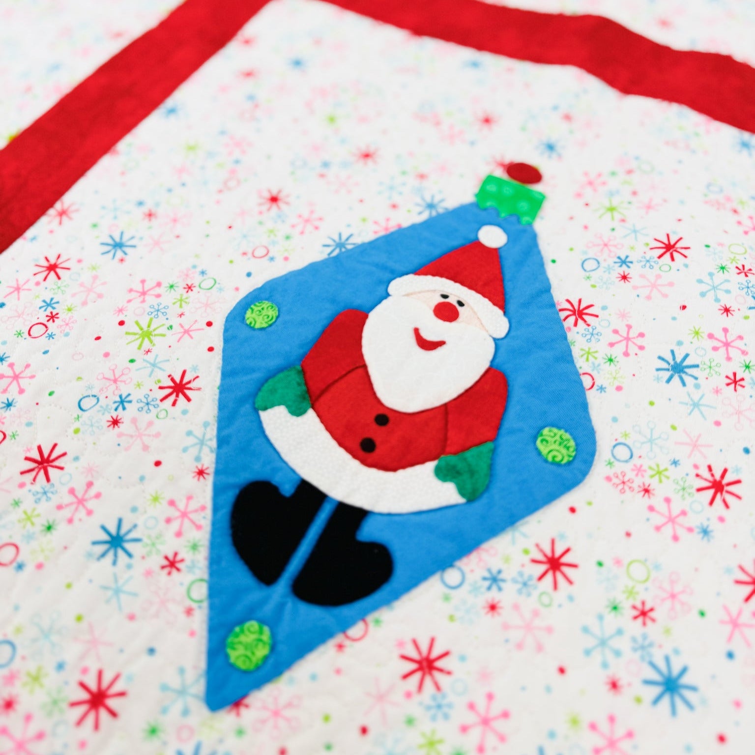 A close-up of a handmade quilt featuring a blue Christmas ornament patch with a smiling Santa Claus design, set against a white quilted background with red ribbon-like stripes and colorful snowflake patterns.