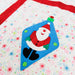 A close-up of a handmade quilt featuring a blue Christmas ornament patch with a smiling Santa Claus design, set against a white quilted background with red ribbon-like stripes and colorful snowflake patterns.