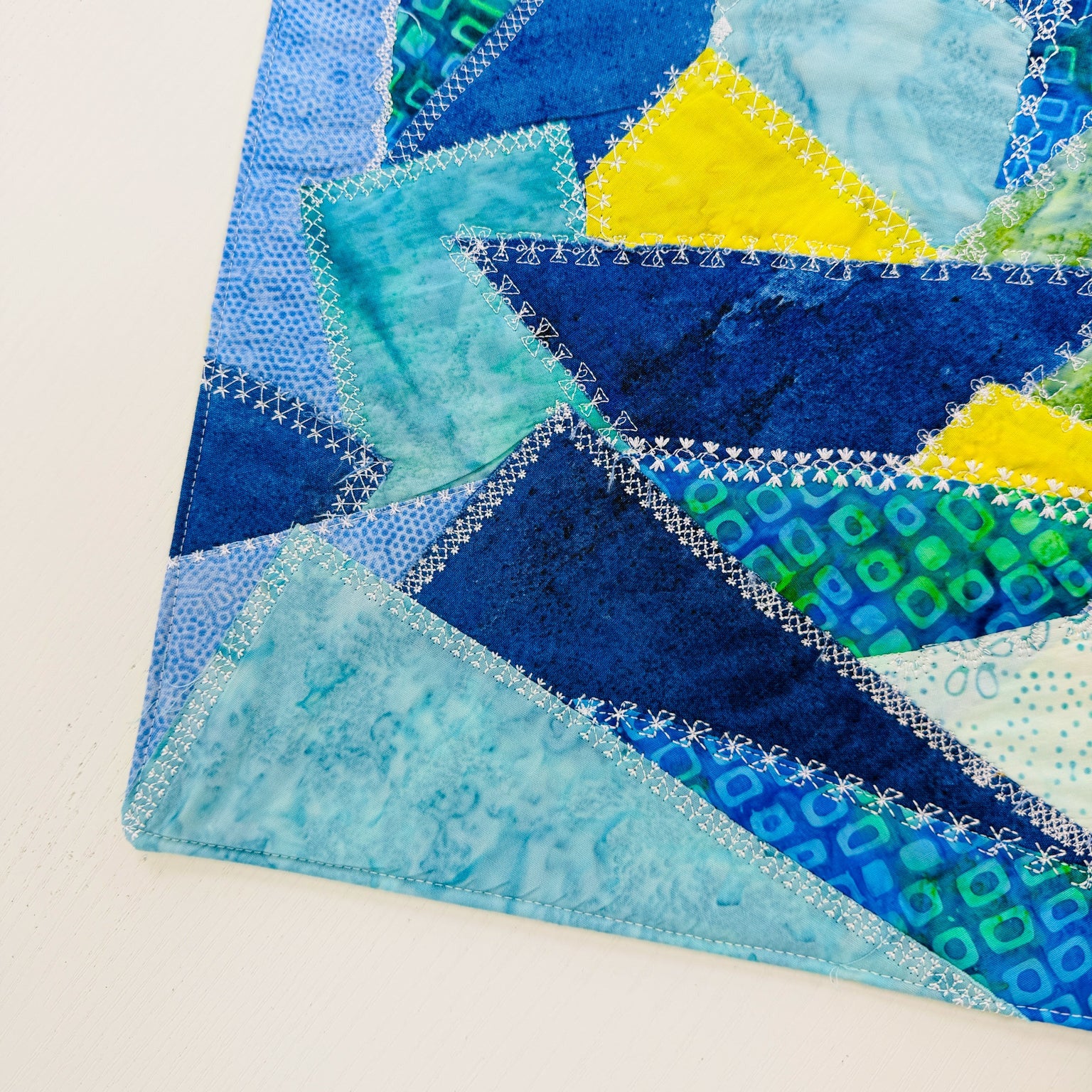 A close-up of a colorful patchwork quilt corner featuring geometric triangles in shades of blue, teal, yellow, and green, with visible white zigzag stitching on a white background.