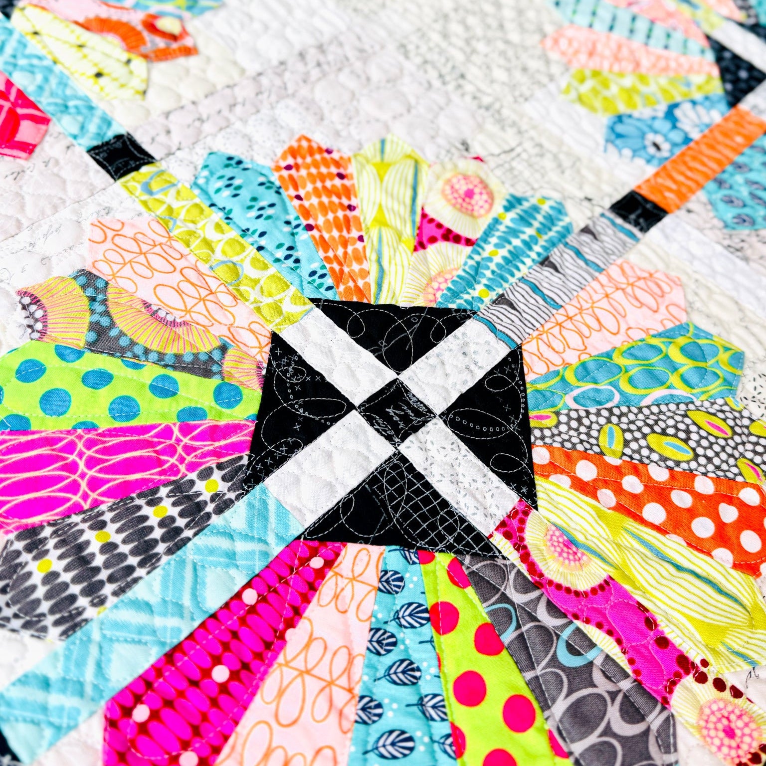 A close-up of a colorful quilted fabric featuring a central black and white checkered square surrounded by brightly patterned radial sections in pink, blue, orange, yellow, green, and gray on a white quilted background.