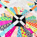 A close-up of a colorful quilted fabric featuring a central black and white checkered square surrounded by brightly patterned radial sections in pink, blue, orange, yellow, green, and gray on a white quilted background.