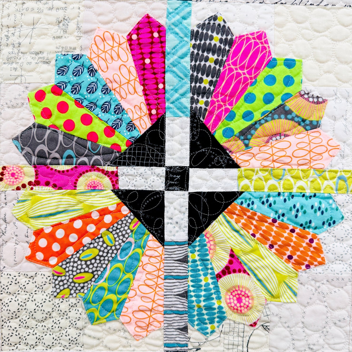 A close-up of a colorful quilted fabric design featuring a symmetrical cross shape made of multiple patterned fabric sections in bright neon, orange, pink, yellow, blue, green, black, and white.