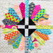 A close-up of a colorful quilted fabric design featuring a symmetrical cross shape made of multiple patterned fabric sections in bright neon, orange, pink, yellow, blue, green, black, and white.