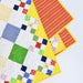 A close-up of a colorful quilted fabric with patchwork squares in white, blue, green, red, yellow, and orange, layered over a bright yellow fabric with red and white stripes, set against a light gray background.