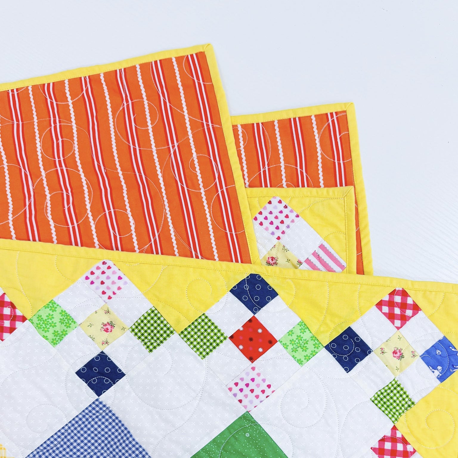 A close-up of a colorful quilted textile with patchwork squares in blue, green, red, pink, and white, layered over bright yellow and orange striped fabric pieces on a light background.