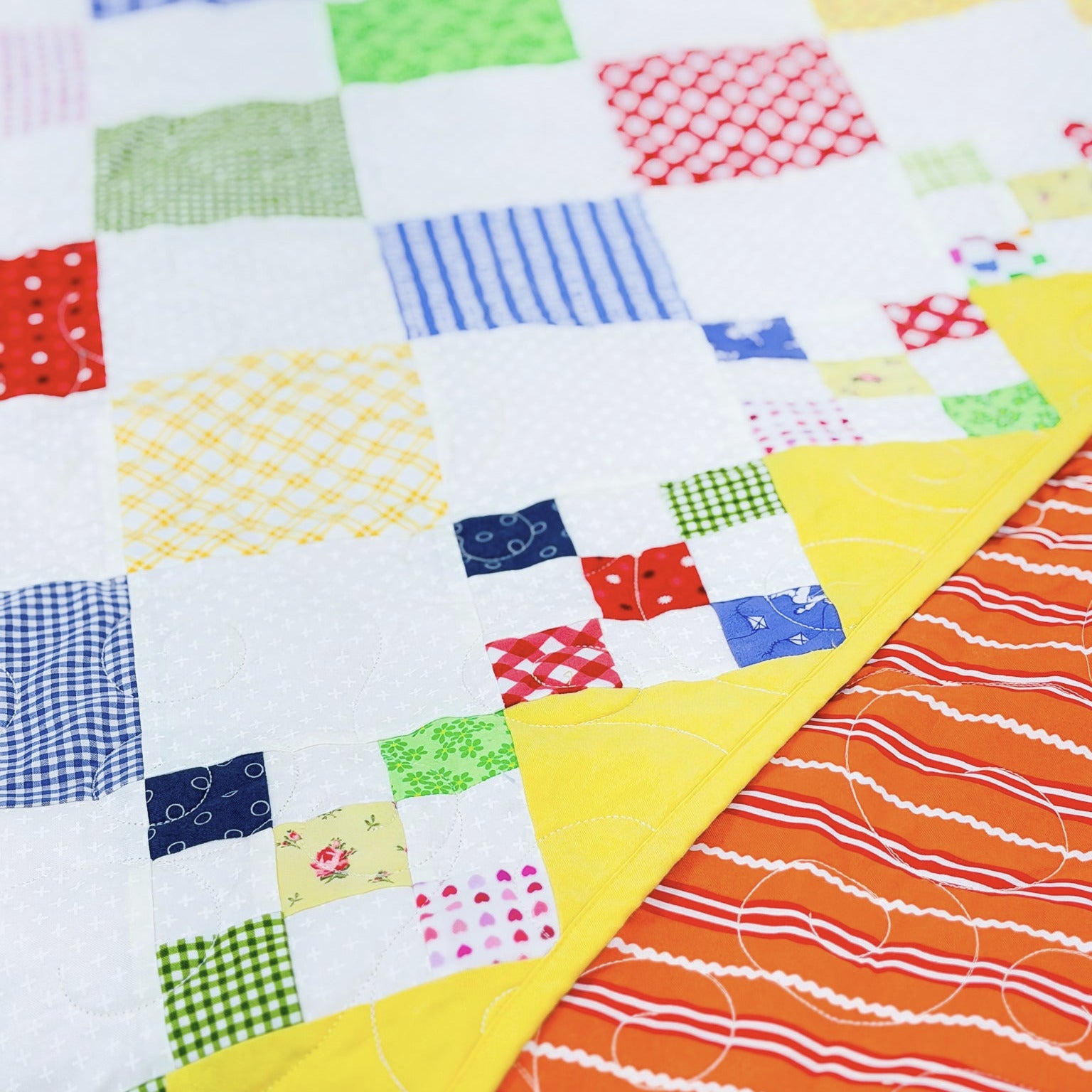 A close-up of a colorful patchwork quilt with white fabric squares, patterned patchwork blocks, and a bright orange quilted section with white wavy lines.