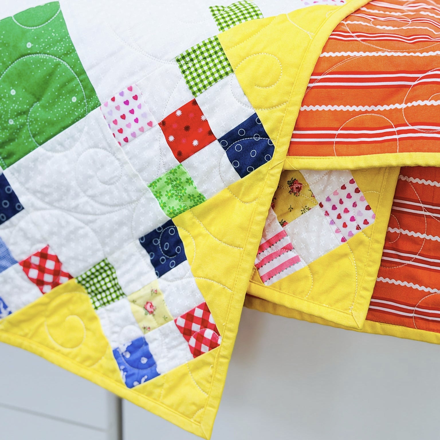 A close-up of a colorful patchwork quilt draped over a white surface, showing yellow borders and multicolored fabric squares including green, red, blue, pink, and orange.