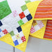 A close-up of a colorful patchwork quilt draped over a white surface, showing yellow borders and multicolored fabric squares including green, red, blue, pink, and orange.