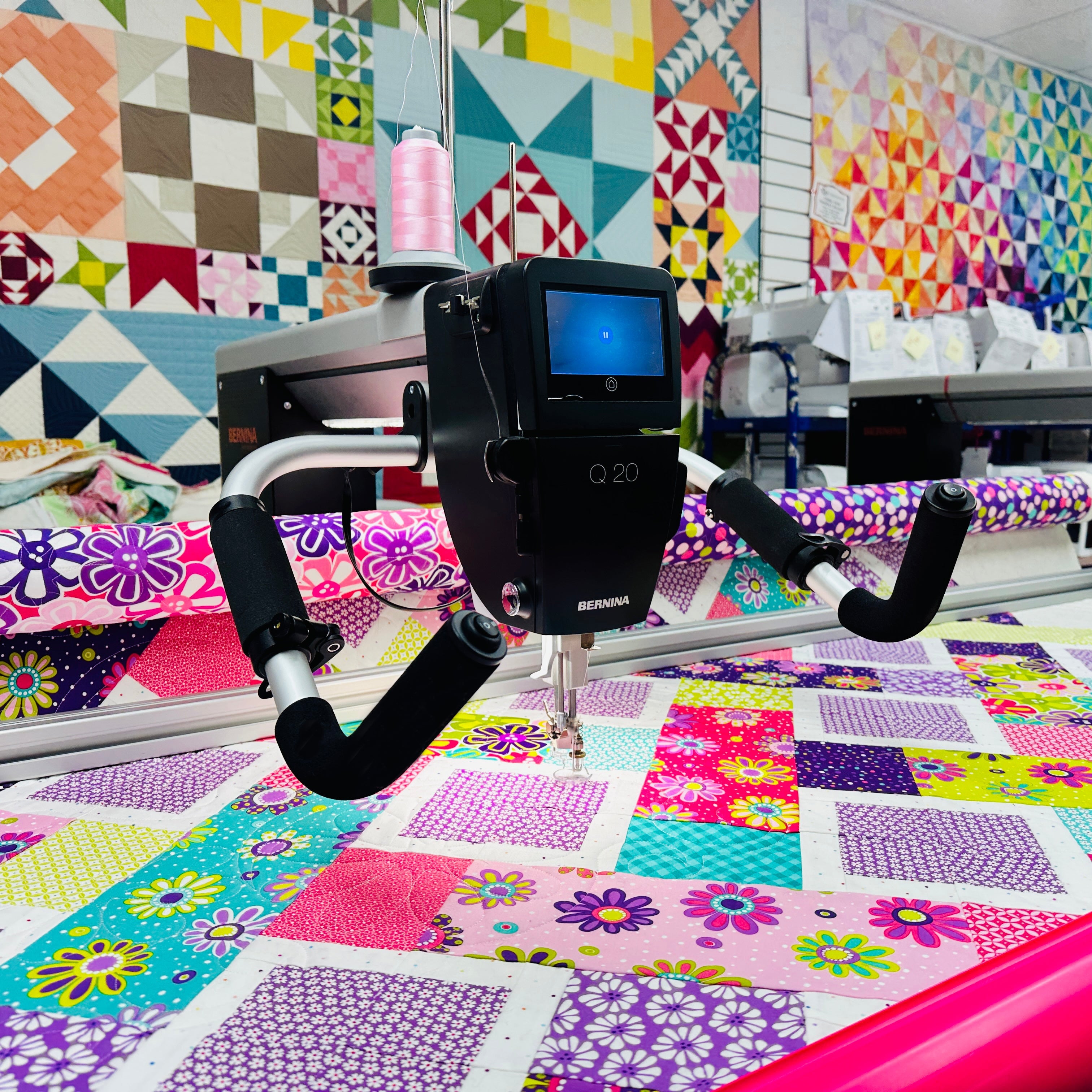 Quilting machine on a colorful quilt with various quilted wall hangings in the background.