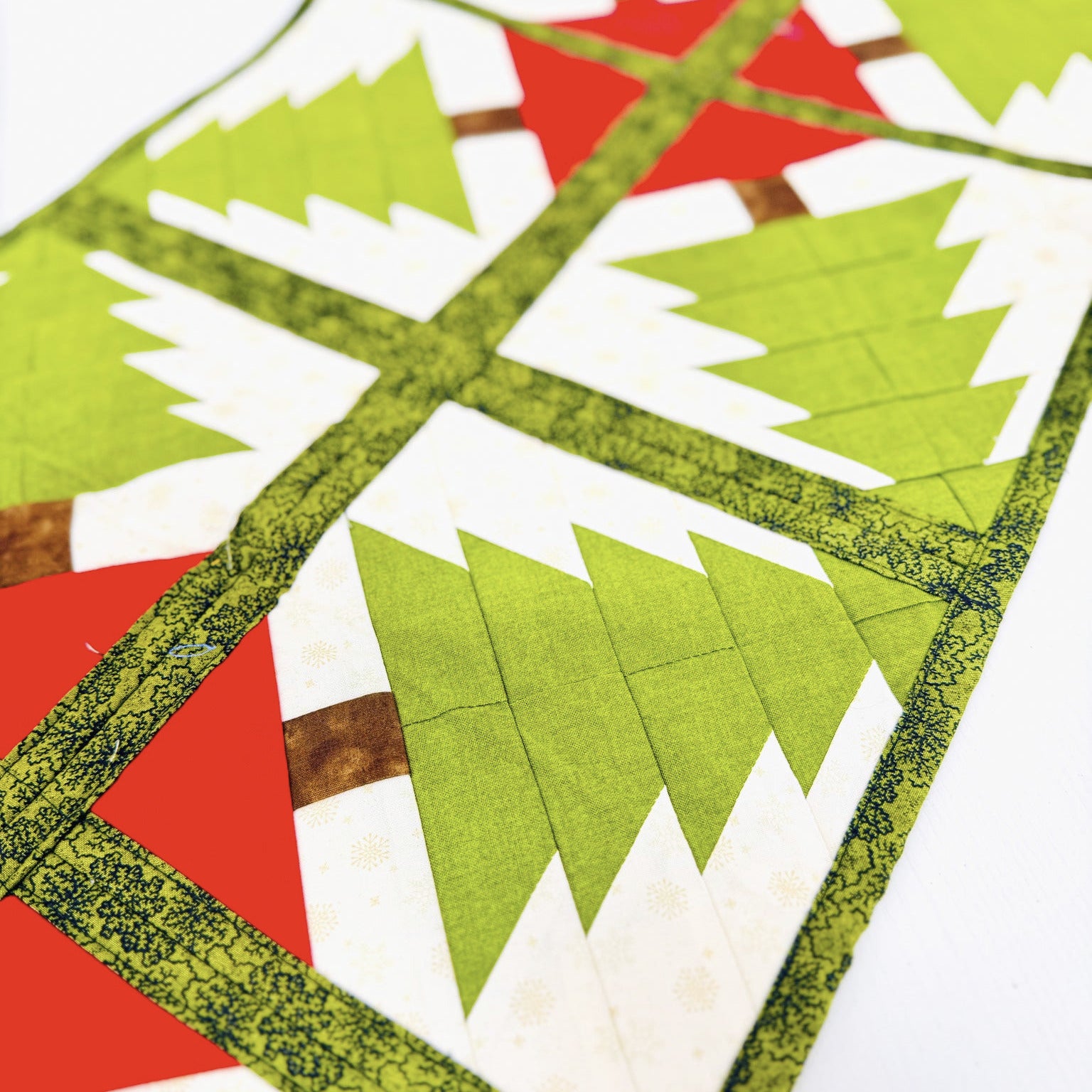 A close-up of a quilted fabric table runner featuring a geometric Christmas tree pattern in green, white, and red, with a decorative olive green patterned border.