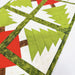 A close-up of a quilted fabric piece featuring a repeating pattern of green Christmas trees on white panels, framed by olive green patterned borders with red and brown accents, laid flat on a white background.