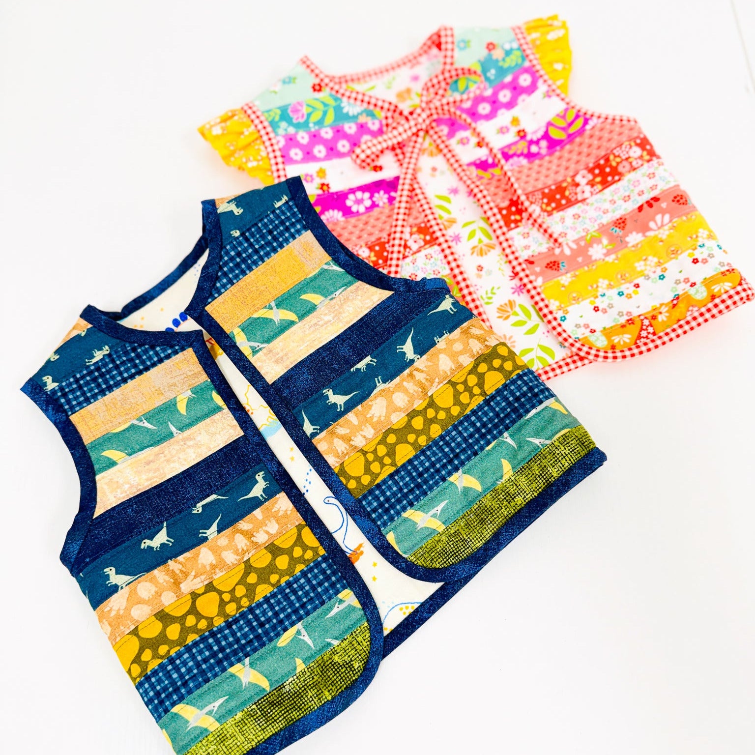 Two sleeveless patchwork vests laid flat on a white background. The front vest is navy blue with horizontal stripes of yellow, green, and orange, featuring a shark pattern.