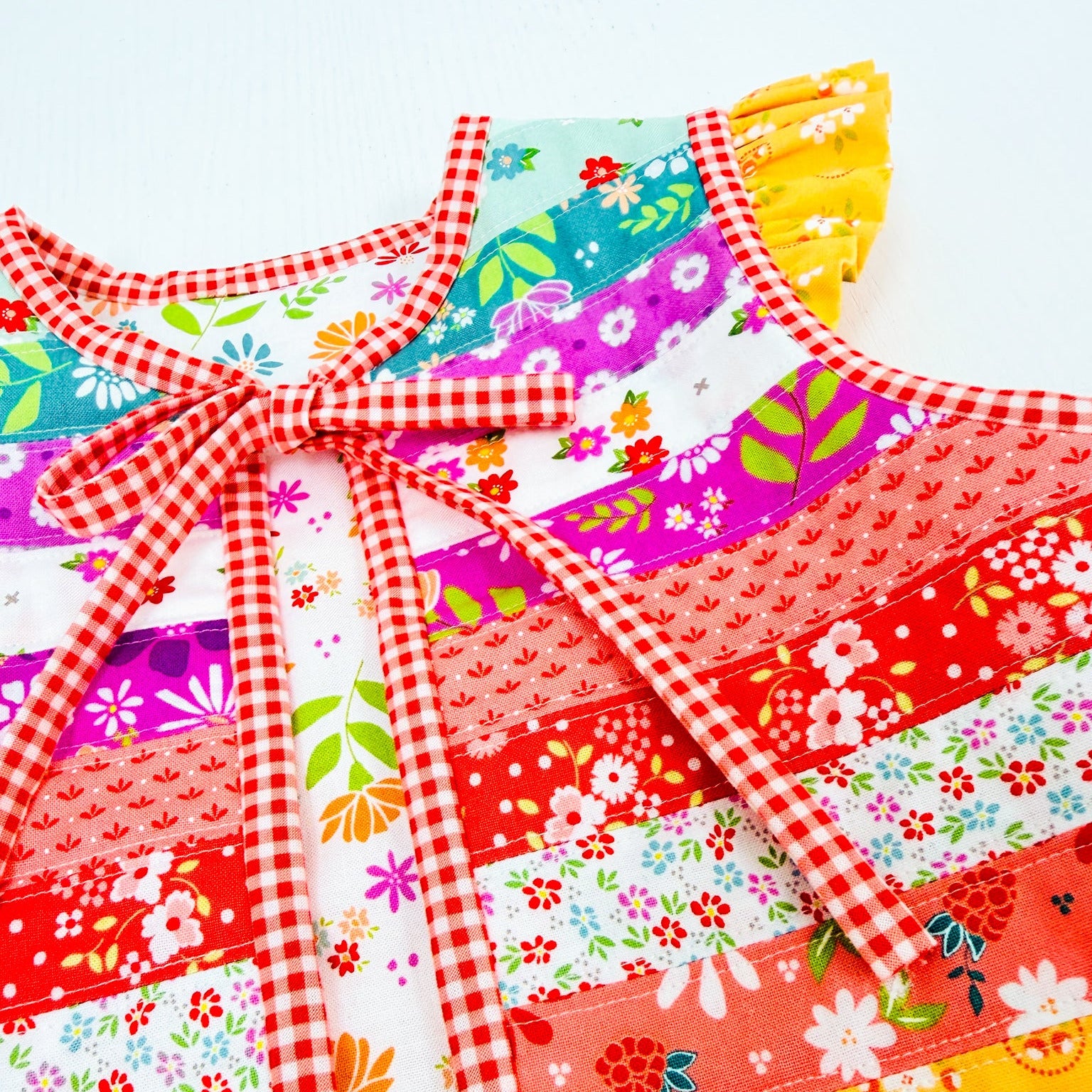 A close-up of a colorful patchwork dress with red gingham trim and a bow, featuring various floral and textured fabric panels in red, yellow, pink, and white.