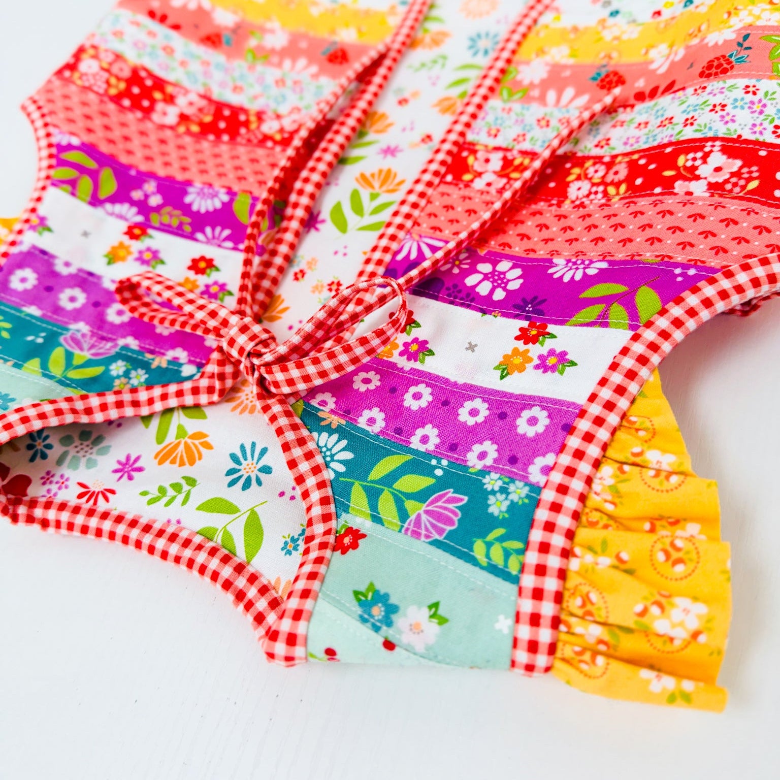A colorful patchwork apron with a red and white gingham border and tie, featuring various floral and patterned fabric panels in pink, purple, blue, yellow, and red, laid flat on a white background.