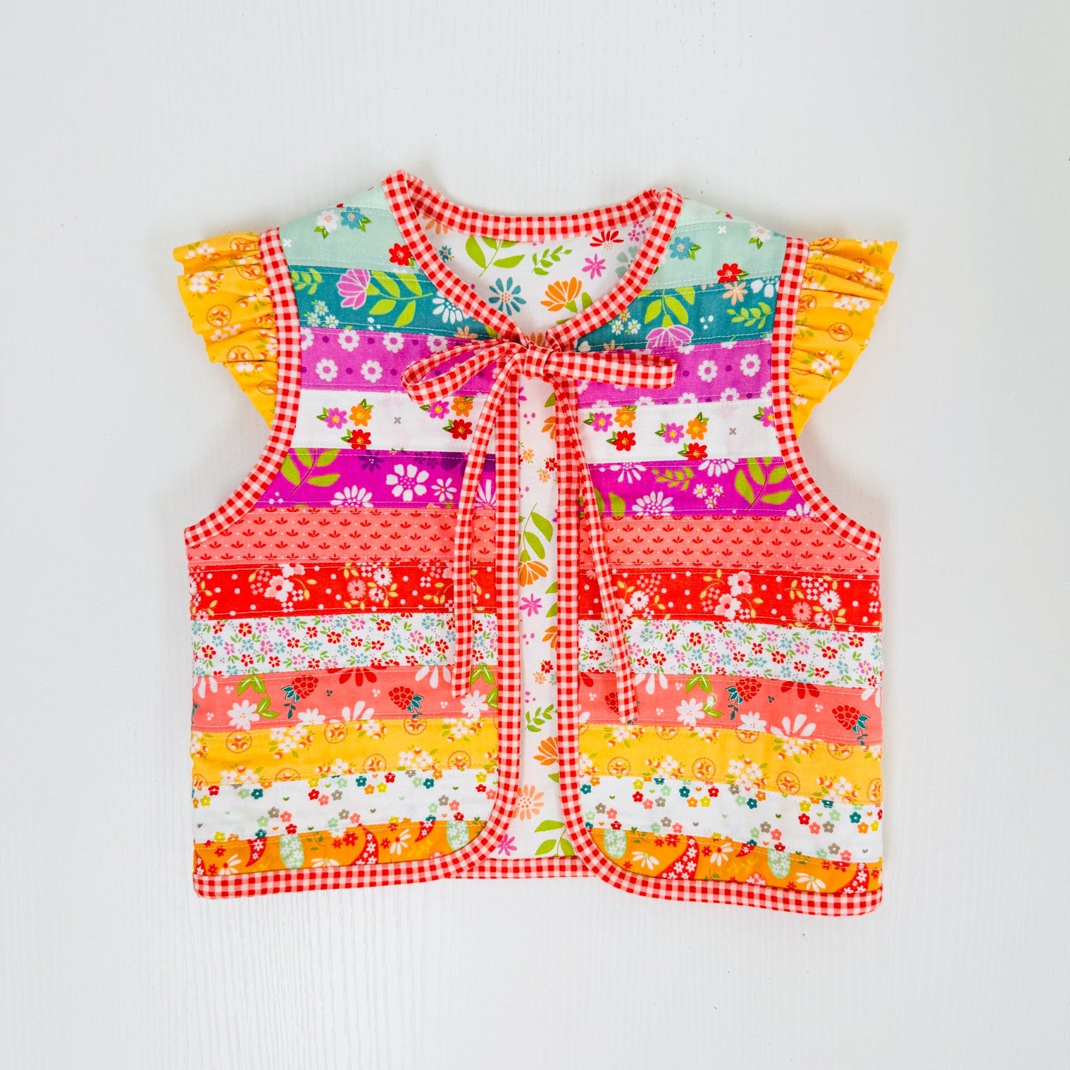 A colorful baby vest with a patchwork floral print, red gingham trim, ruffled sleeves, and a front tie closure, laid flat on a white background.