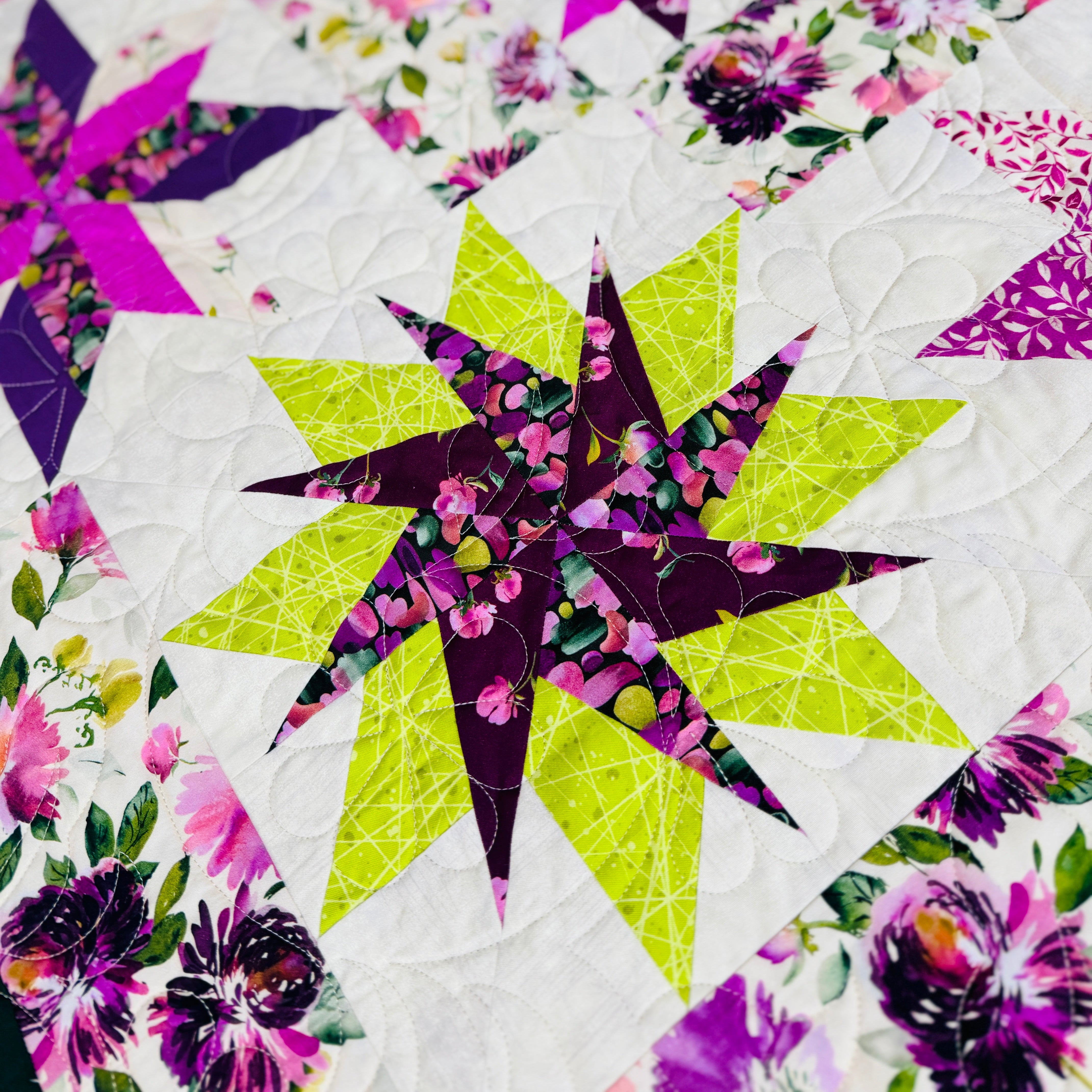 A close-up of a colorful quilted fabric featuring geometric star and floral block patterns in purple, pink, lime green, and white.