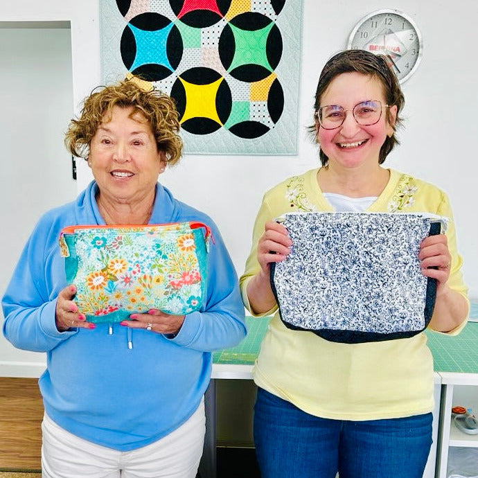 Two women holding fabric pouches in a room with a colorful quilt in the background.