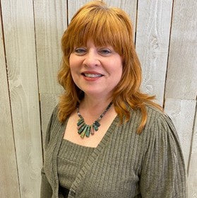 Woman with red hair wearing a green sweater and necklace against a wooden paneled wall.
