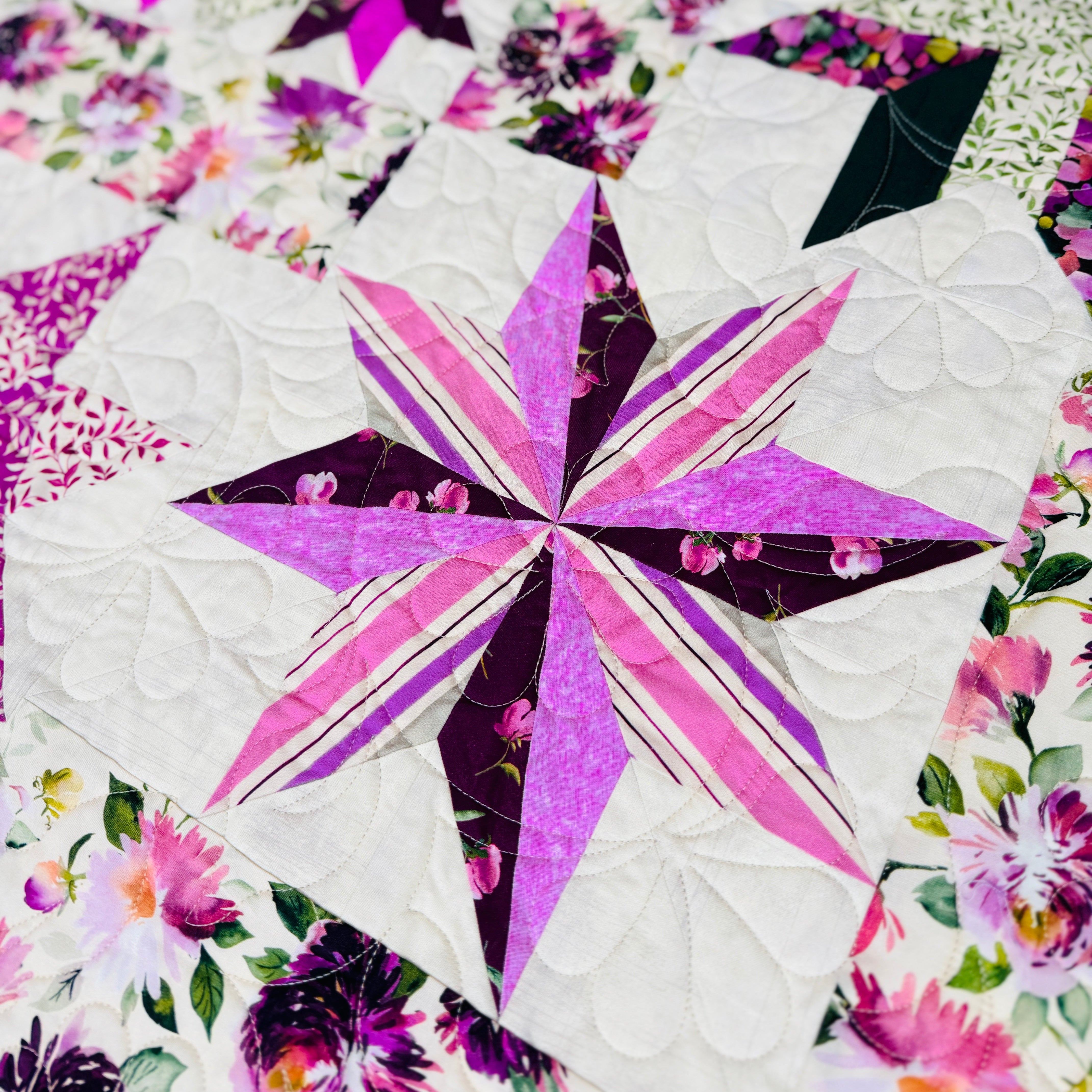 A close-up of a quilted fabric featuring a starburst patchwork design in shades of pink, purple, and white, surrounded by floral patterns.