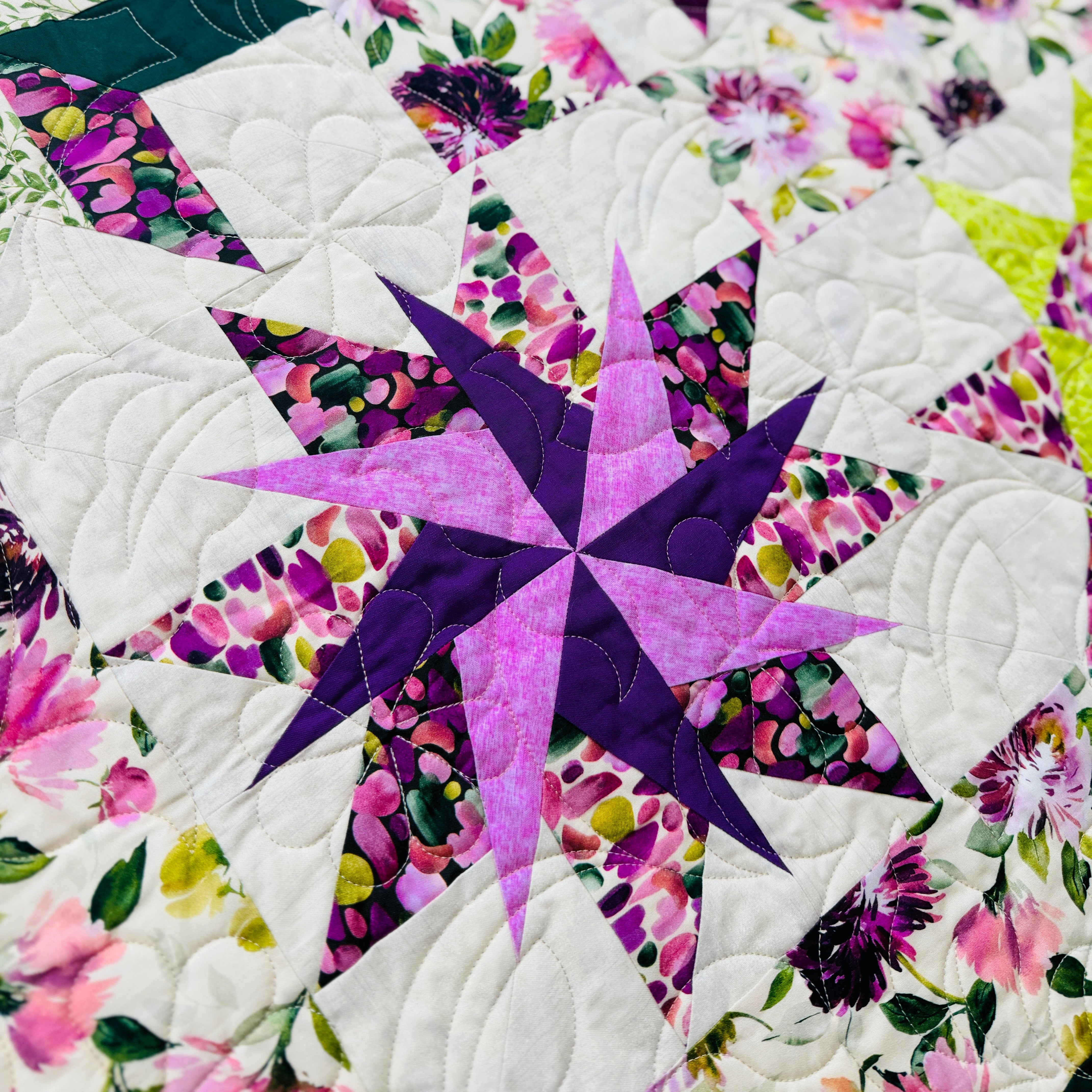 A close-up of a quilted fabric featuring a geometric star pattern in purple and pink, surrounded by white quilted sections and floral prints with green leaves and pink flowers.