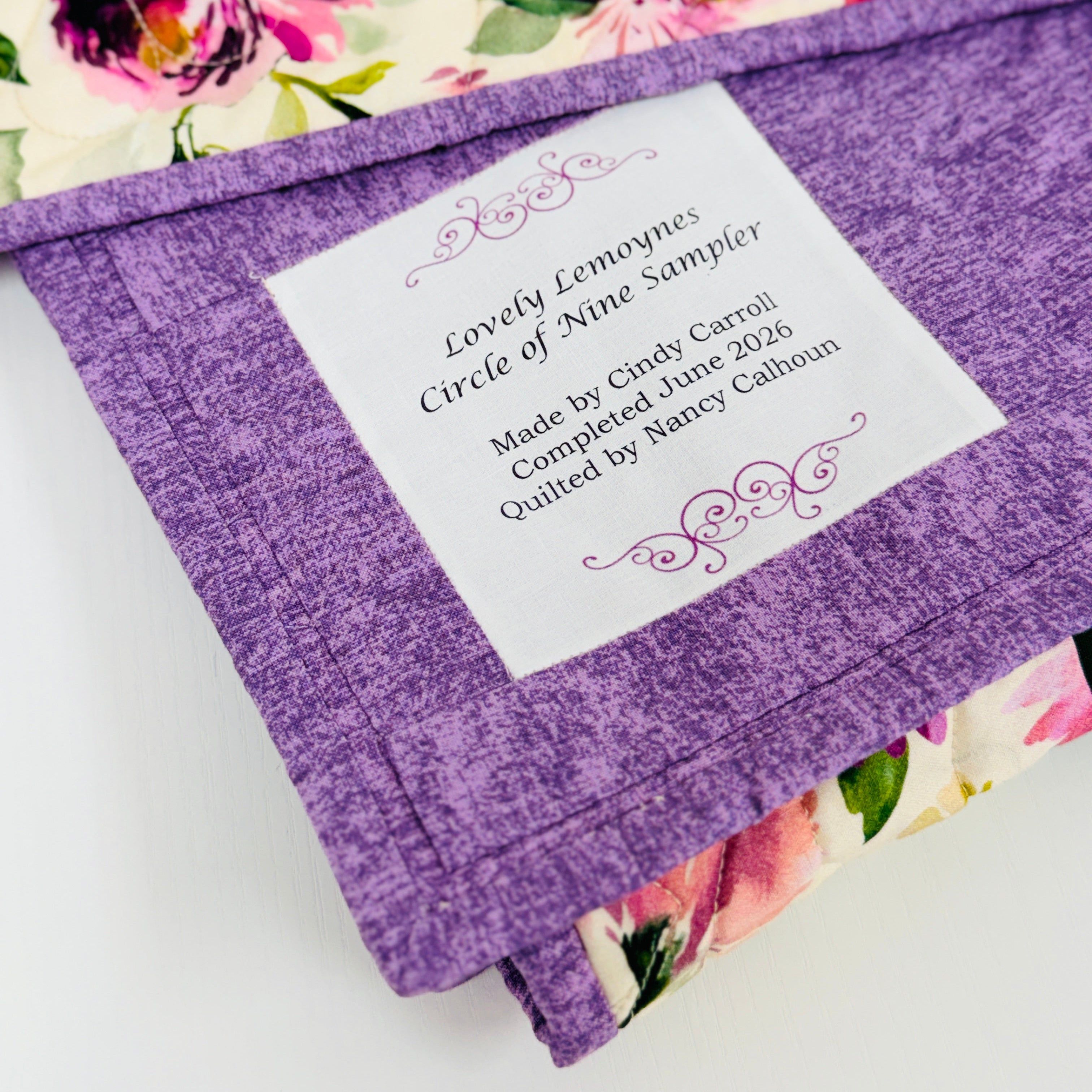 A close-up of a purple fabric envelope-style label holder with a white paper insert showing printed text, placed over a floral quilted fabric background.