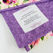 A close-up of a purple fabric envelope-style label holder with a white paper insert showing printed text, placed over a floral quilted fabric background.
