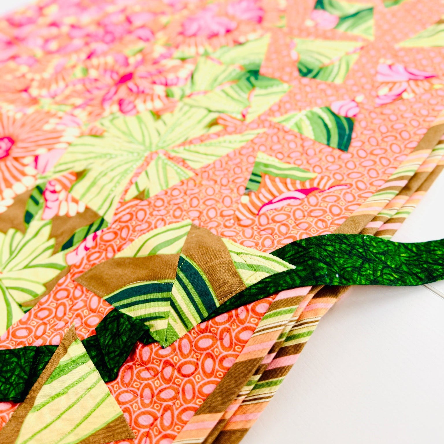 A close-up of a colorful quilted fabric item, likely a quilt or textile panel, featuring a patchwork design with pink floral and green tropical leaf patterns on a coral background, with a dark green ribbon tied around it.