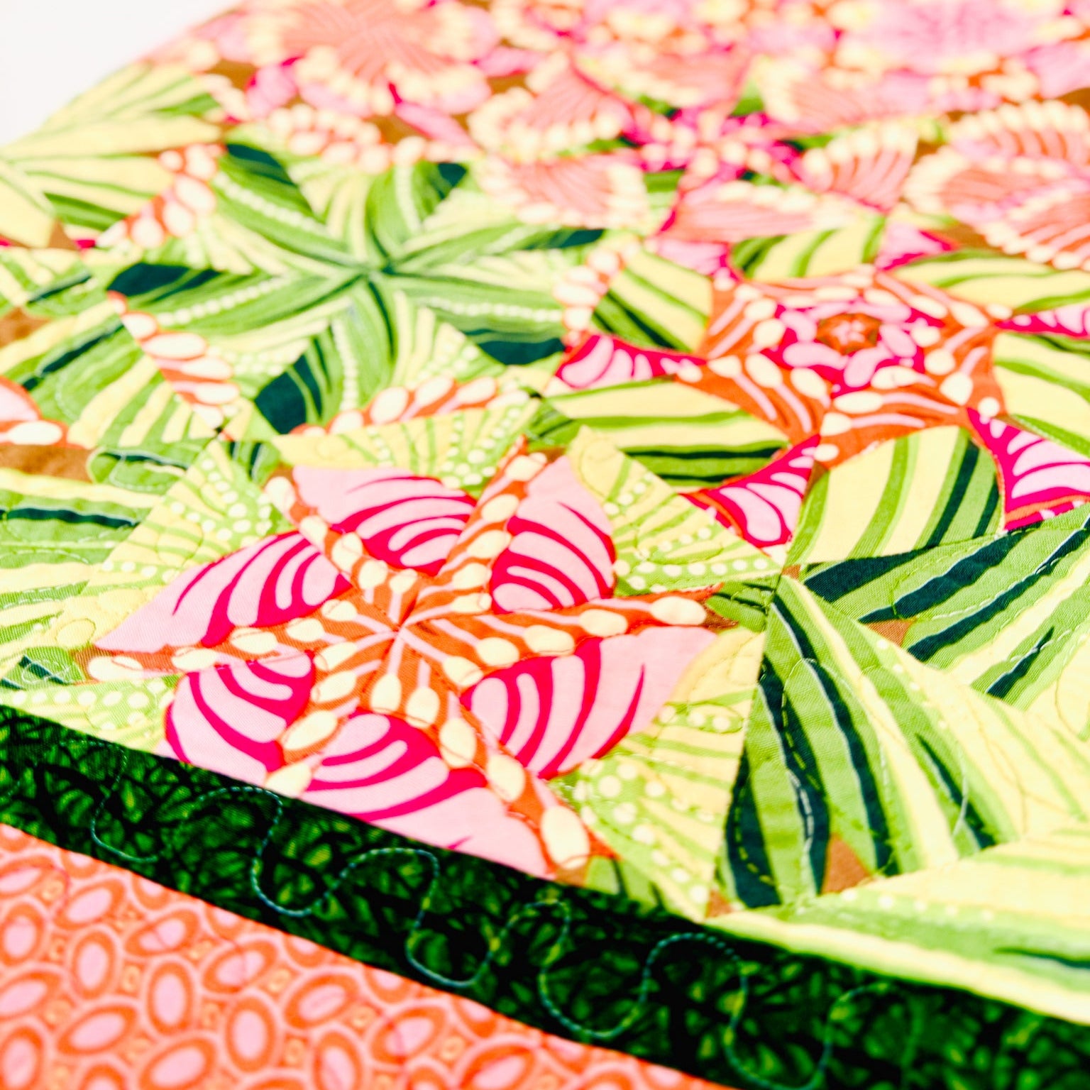 A close-up of a colorful quilted fabric featuring a tropical floral and leaf pattern in shades of green, pink, orange, and yellow, with a dark green stitched border.