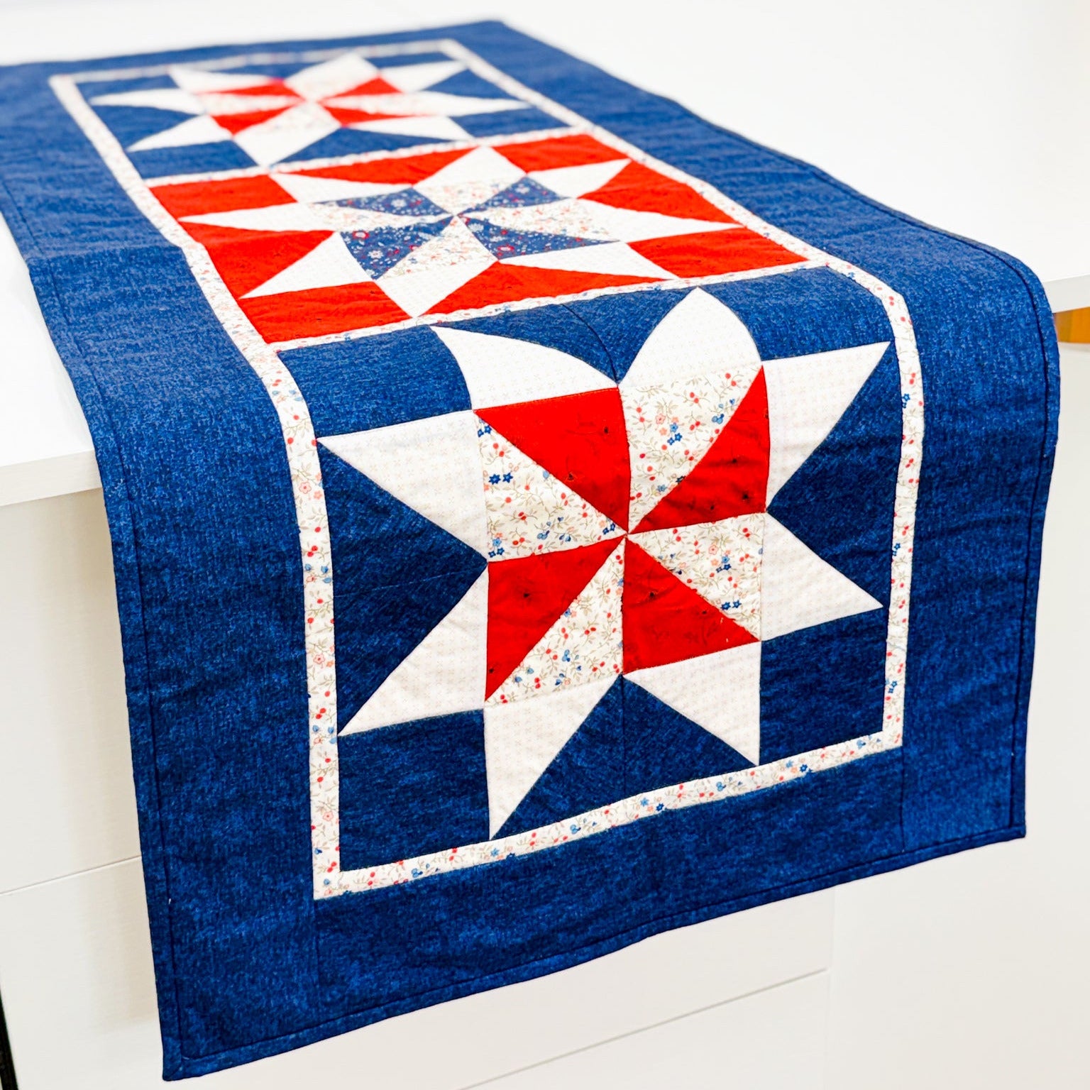 A rectangular quilted table runner with a red, white, and blue patriotic starburst pattern draped over a white table against a white wall.