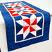 A rectangular quilted table runner with a red, white, and blue patriotic starburst pattern draped over a white table against a white wall.