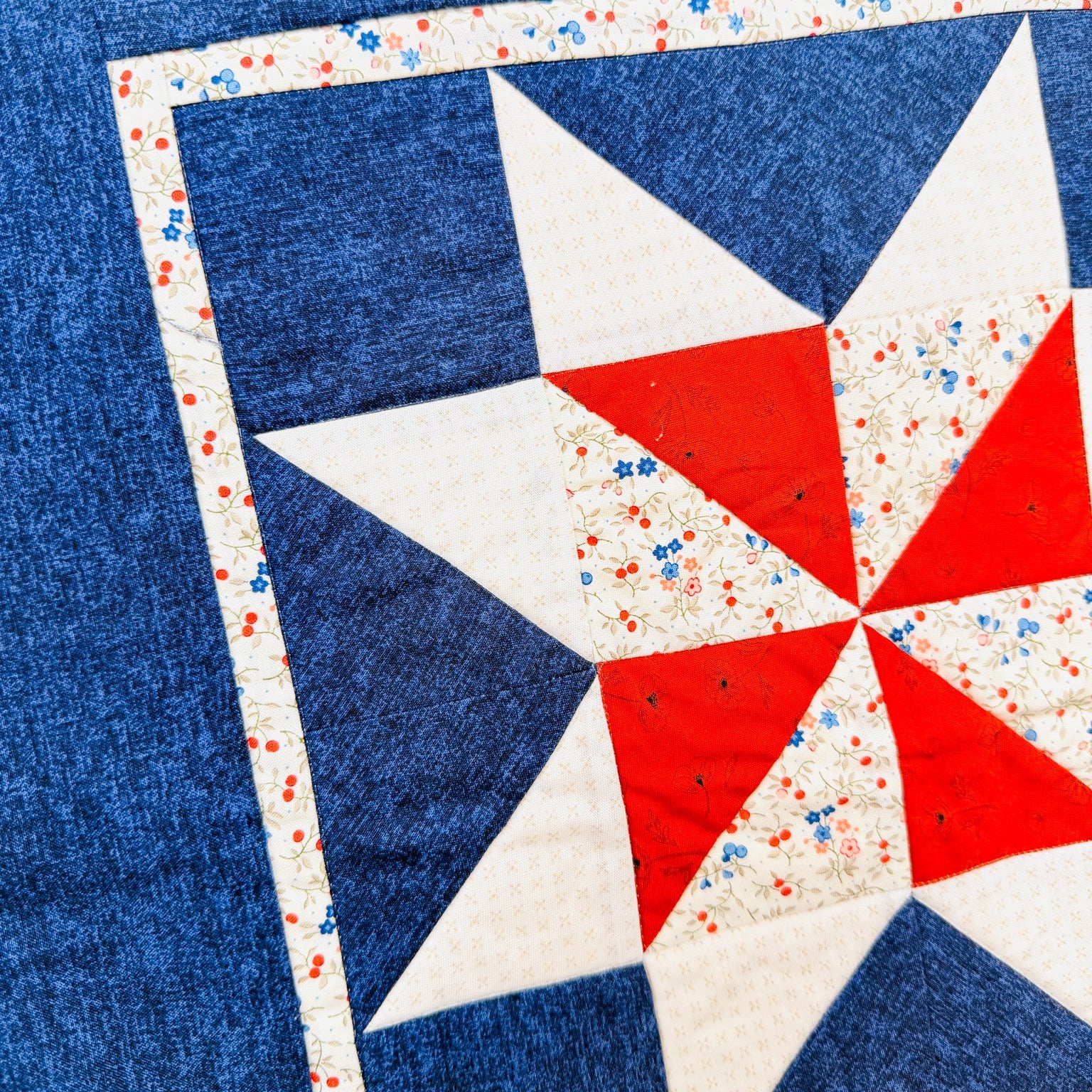 Close-up of a quilted fabric featuring a geometric star pattern with blue, white, and red sections, bordered by a white fabric with small red and blue speckles.
