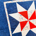 Close-up of a quilted fabric featuring a geometric star pattern with blue, white, and red sections, bordered by a white fabric with small red and blue speckles.