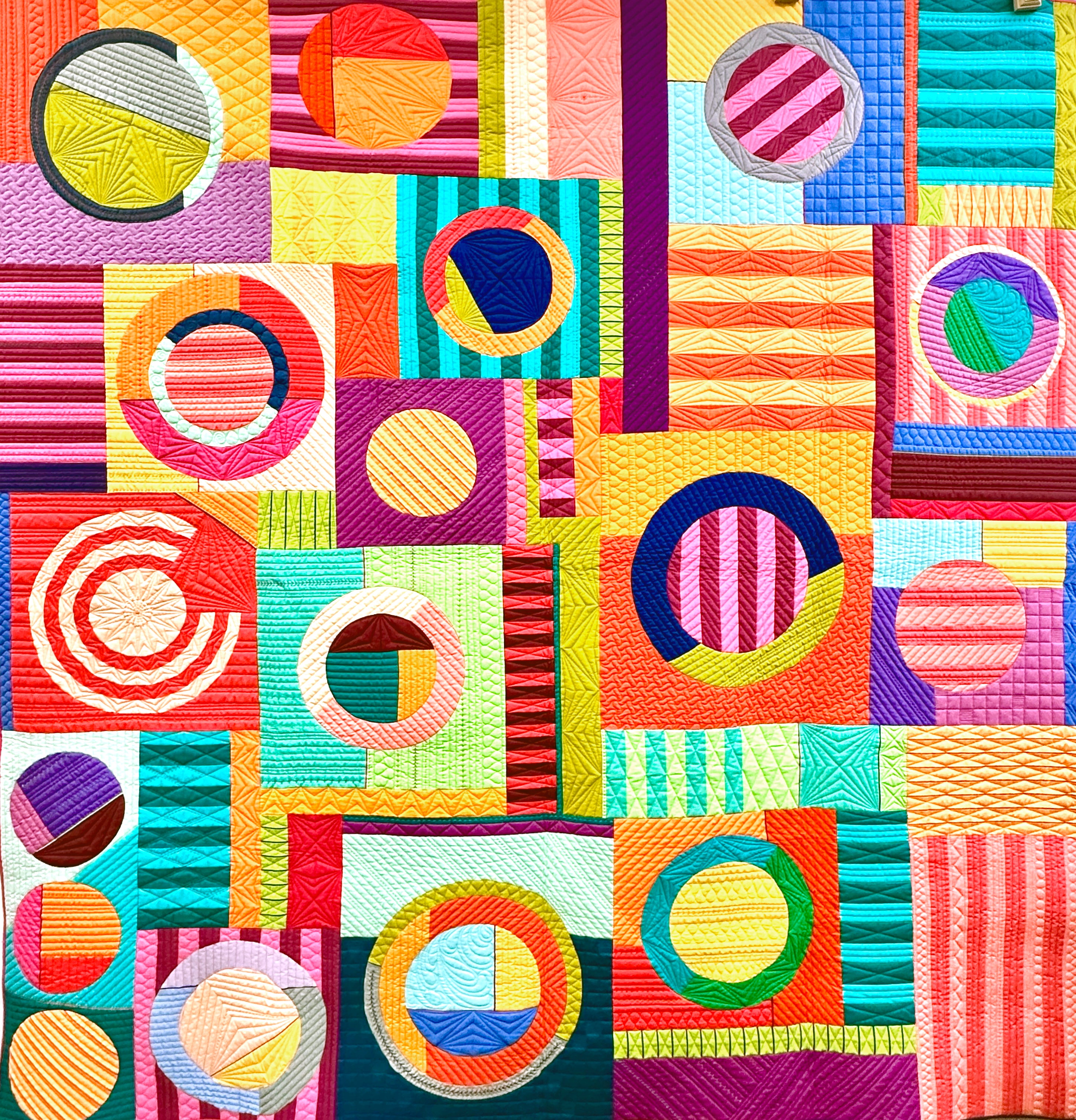 A vibrant, multicolored geometric patchwork quilt featuring circular, striped, and solid patterned blocks in bright shades of pink, yellow, blue, green, orange, and purple.