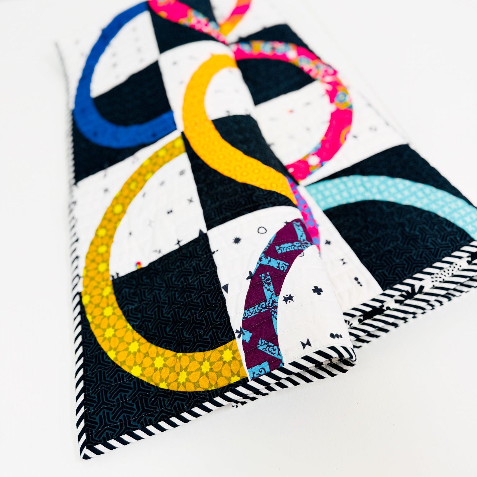 A folded quilted fabric item, likely a small pillow cover or pouch, featuring a modern abstract design with black and white checkered squares and curved stripes in yellow, blue, pink, and light blue, photographed on a clean white background.