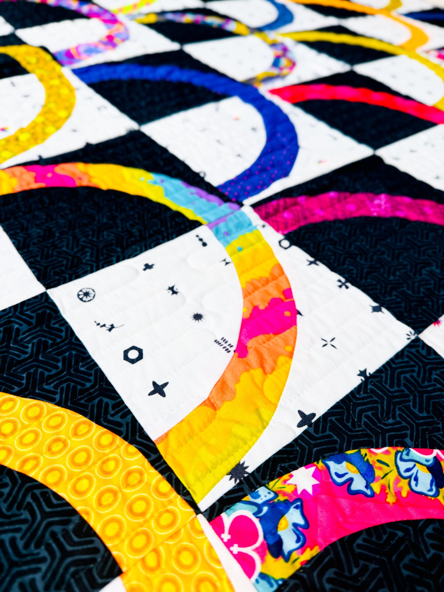 A close-up of a colorful quilted fabric with geometric and rainbow patterned sections in black, white, yellow, blue, pink, and orange.