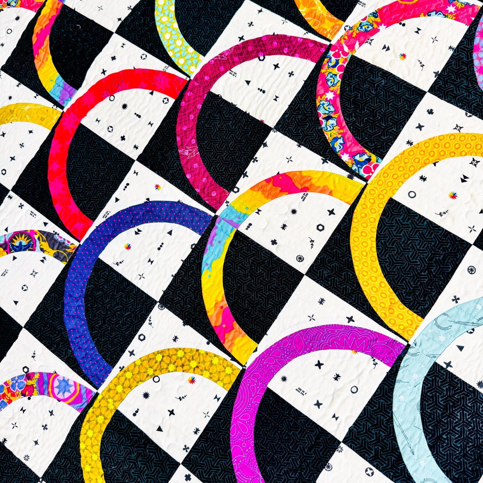 A close-up of a colorful quilted fabric pattern featuring alternating black and white checkered squares with curved semicircle arcs in bright pink, yellow, blue, purple, green, and orange, some with floral or textured prints.