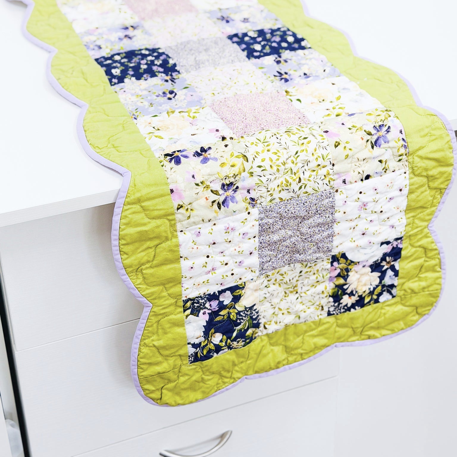 A quilted table runner with a scalloped edge, featuring a patchwork design of floral and solid fabric squares in yellow, navy, pink, and white, with a bright yellow-green border and light purple trim, displayed on a white table.