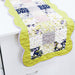 A quilted table runner with a scalloped edge, featuring a patchwork design of floral and solid fabric squares in yellow, navy, pink, and white, with a bright yellow-green border and light purple trim, displayed on a white table.