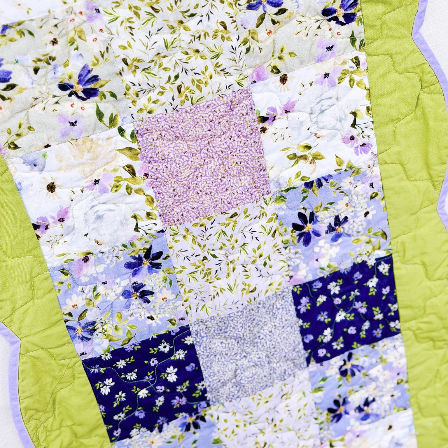 A close-up of a handmade patchwork quilt featuring various floral and patterned fabric squares in pastel shades of blue, pink, purple, and white, bordered by a bright lime green quilted edge.