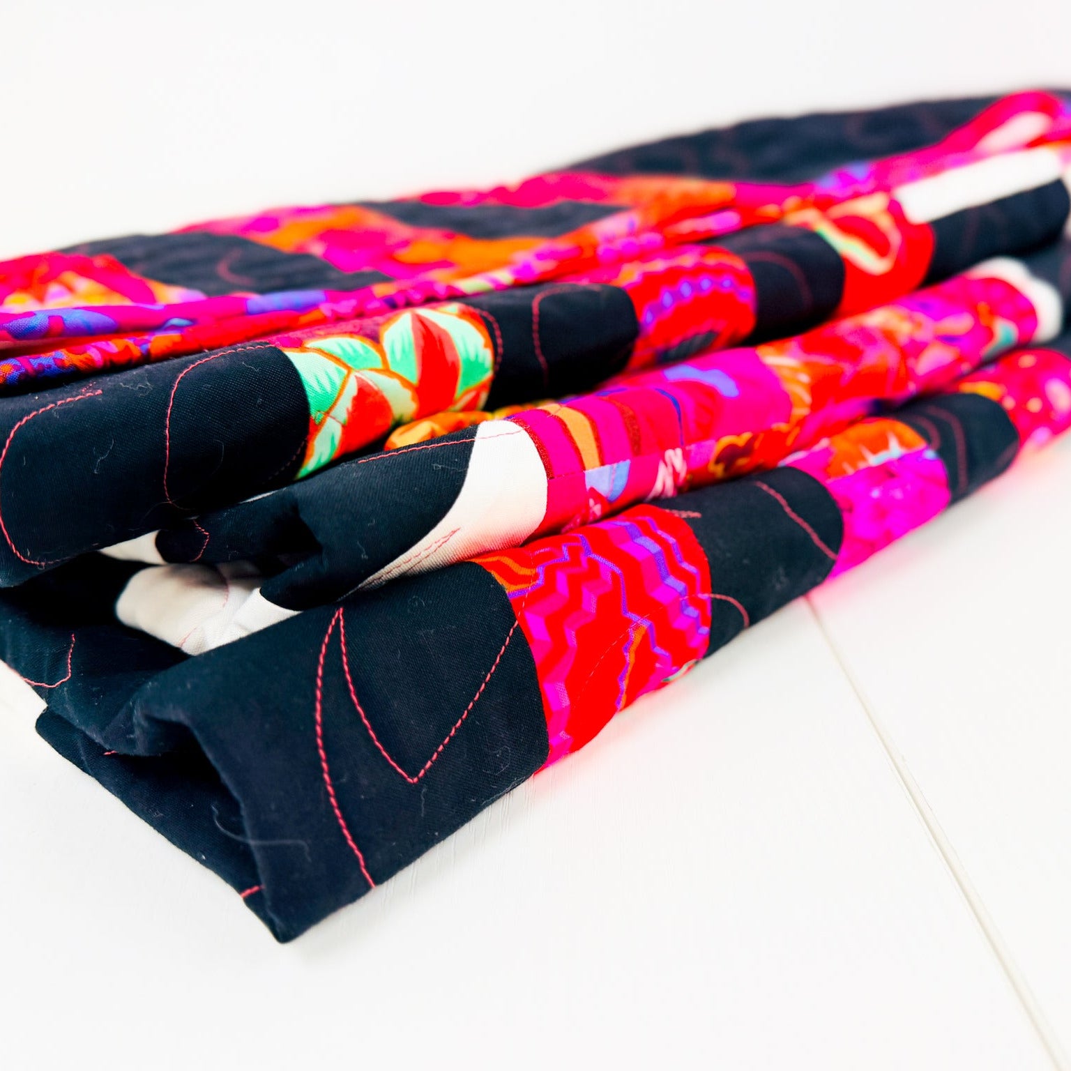 A folded quilted textile with a bold multicolor pattern, featuring black sections and bright pink, red, orange, green, and white abstract designs, photographed on a clean white surface.