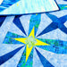 A close-up of a quilted textile with a geometric starburst pattern in shades of blue, teal, and green, featuring a bright yellow star center and a folded section showing the reverse side.